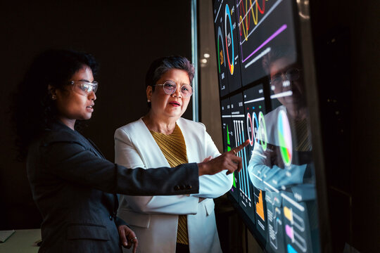 Two professionals collaborate at a large interactive screen, pointing at colorful charts and graphs on a data dashboard, analyzing metrics and strategy in a modern office