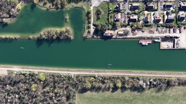 Aerial view of a straight canal reflecting sunlight, flanked by green trees, houses, and docked boats, creating a vibrant tableau, Sausheim, Grand Est, France.