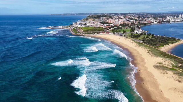 Aerial drone video flying over Nobbys Beach with waves crashing onto the sandy shoreline, moving towards the beach pavilion near the harbour entrance in Newcastle, New South Wales, Australia.