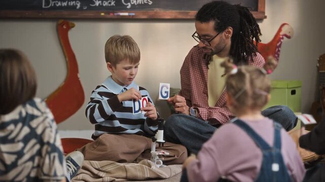 Full shot of young biracial male teacher helping Caucasian boy to spell word dog using flashcards during group educational activity in kindergarten classroom, sitting on floor in front of blackboard