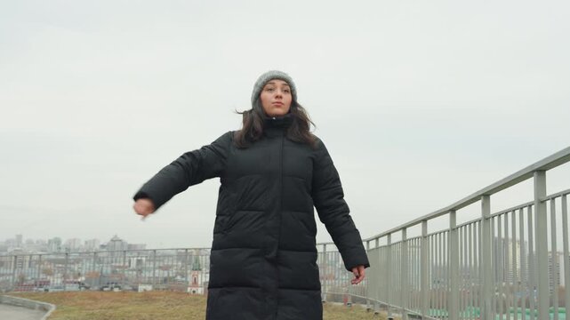 Woman dancing on city hilltop bundled in black coat and beanie, improvising expressive arm gestures along railing, alternating wide steps and close portraits, overcast skyline backdrop, cold breath