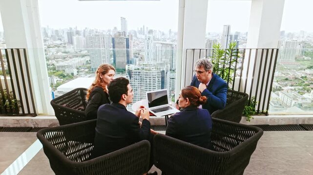 Four professionals meet on a rooftop terrace with a laptop, discussing work against a modern city skyline.