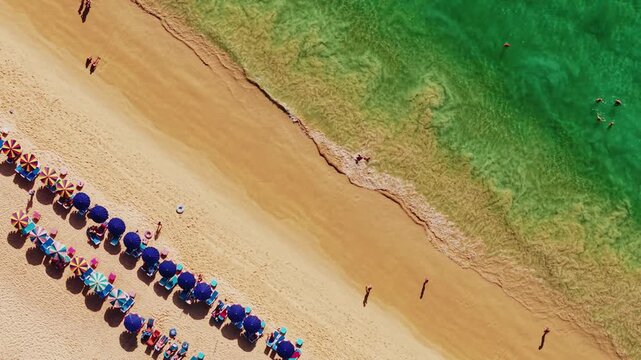 Aerial tracking view of beach umbrellas and people along sandy shore with rolling waves, Karon on Phuket island, Thailand.