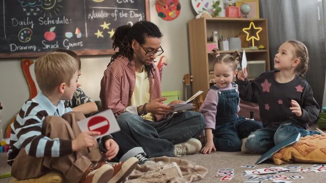 Full slowmo shot of focused young biracial kindergarten worker teaching road signs to playful Caucasian children, sitting on floor in modern classroom and handing out flashcards to everyone