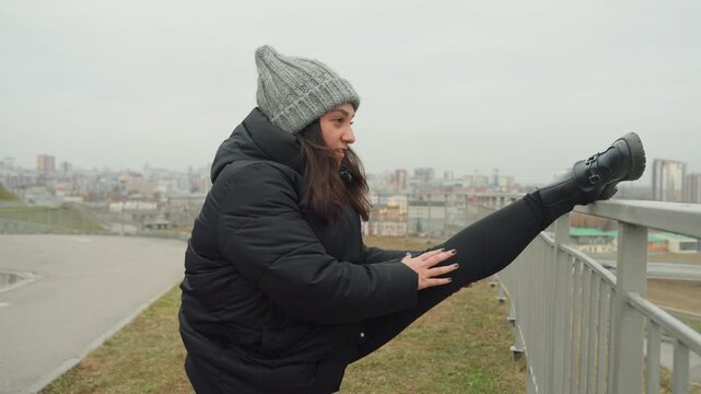White woman performing high kick outdoors, winter coat and knit beanie, city skyline backdrop, upbeat dance warmup and playful rehearsal, arm rhythm and strong balance, urban overlook choreography
