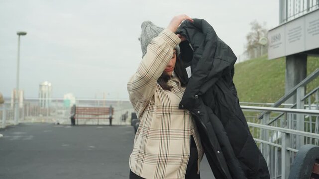 Closeup woman removing black jacket on rooftop, knit beanie and patterned sweater, adjusting collar and hood, candid urban movement, cool overcast mood, bench and railing in background