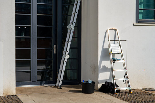 Aluminum multi-purpose ladder and stepladder leaning against a white modern building wall next to a black bucket, ready for window cleaning, renovation works, or professional exterior facility mainten