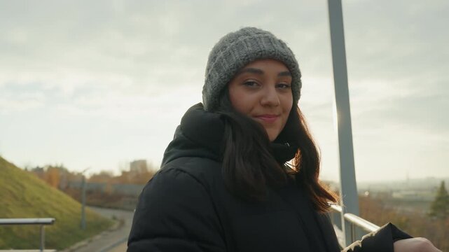 Young woman rooftop sunrise gentle dance. Outdoor overlook with metal railing and grassy hill, knit beanie and puffer jacket catching golden backlight. Soft turns and shy smile, lens flare