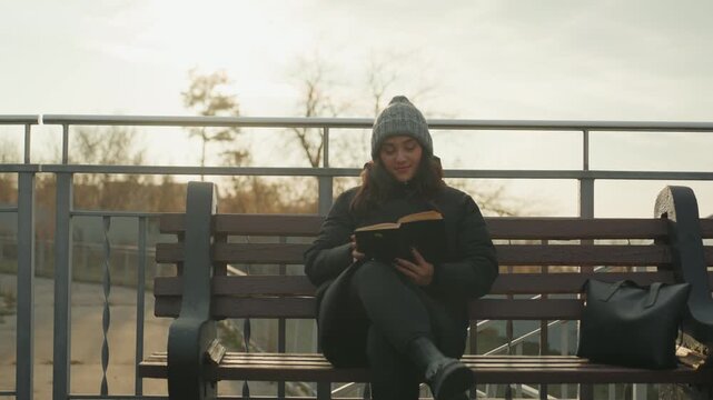 Woman reading on park bench, golden backlight and urban railing. Studentstyle pause between classes, beanie and winter coat, backpack at side, pages turning slowly, soft autumn light, calm