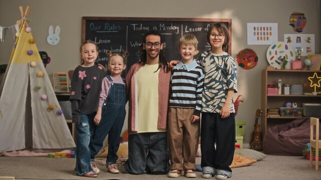 Full slowmo portrait of young biracial male kindergarten teacher smiling at camera with cheerful Caucasian children, posing together in cozy classroom in front of chalkboard and colorful teepee tent