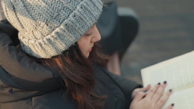 Woman flipping pages on book, midshot of reader in knit beanie and puffer jacket turning worn pages, closeup on hands and nails, urban sidewalk setting, soft evening light, calm deliberate rhythm