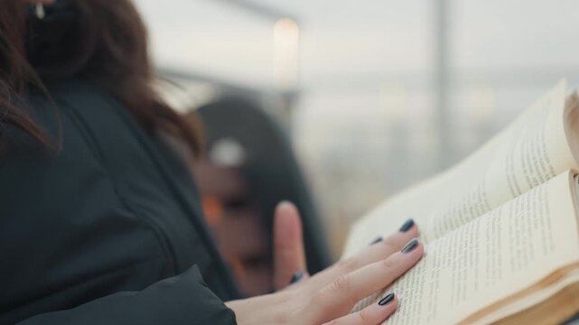 Closeup hands reading worn book, soft finger movements turning yellowed pages, black nail polish, dark coat, blurred city skyline background, slow cinematic pacing, intimate reflective mood, tactile