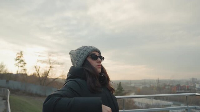 Wide cityscape dancer on balcony framed by metal railings lowangle shots capturing skyline and evening light slow fluid movement empowered stance winter hat and sunglasses atmospheric solitude