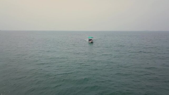 Drone ascending pullback from small boat with turquoise canopy centered in calm Arabian Sea waters off Malvan coast, Maharashtra. Overcast sky creates soft lighting across expanding seascape.