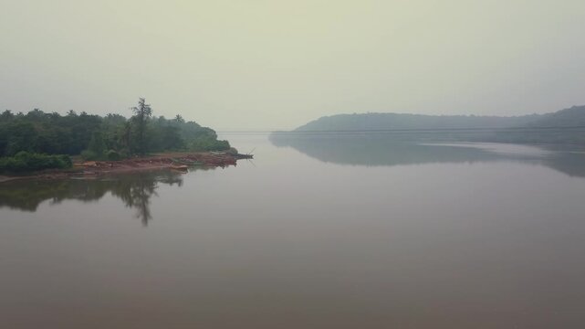 Wide view of the calm Karli River winding between forested banks and low hills near Tarkarli, Malvan in Maharashtra, India. Misty atmospheric conditions with still water reflecting vegetation.