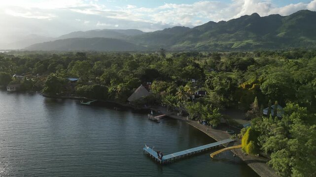 Lake Izabal shoreline in Guatemala. Drone flies along water edge past fincas with low sun casting soft shadows, hazy hills in distance and blue sky with scattered clouds.
