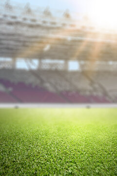 Close-up of lush green grass on a professional soccer field with a blurred stadium and bright sunlight in the background. Ideal for sport-themed designs