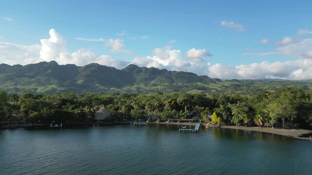 Lake Izabal shoreline in Guatemala. Rising drone shot, fincas along water edge, expanding to farmland and rolling hills in background with lake surface in foreground.