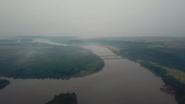 Calm brown river waters flow between forested banks and exposed red soil shores under hazy skies in the Karli River backwaters near Tarkarli and Devbag, Malvan district, Maharashtra, India.