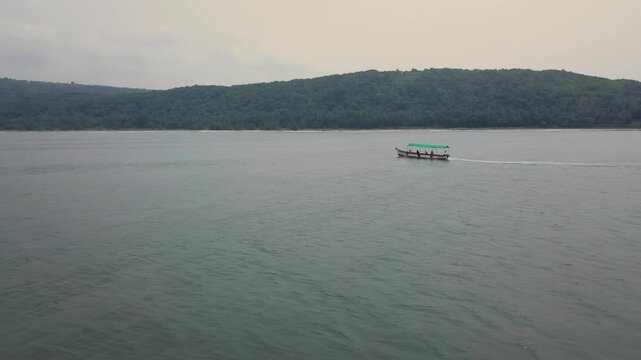Tourist boat moving through tranquil green ocean waters with forested coastline in background, Malvan coastal region, Maharashtra, India during post-monsoon season on overcast day