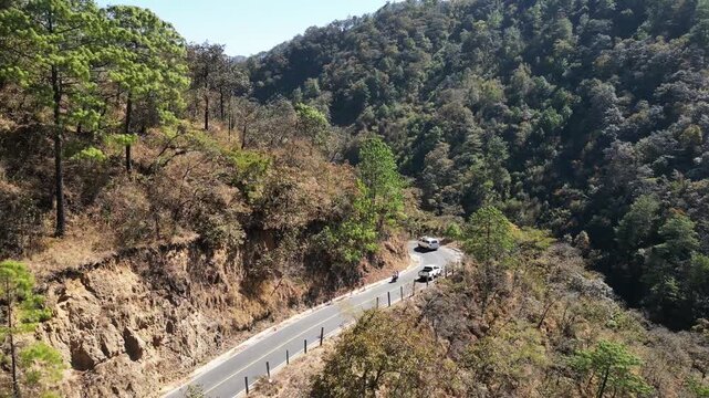 High-angle drone shot of cars driving along a winding paved road in Guatemalan highlands, showing dry hills, valleys.