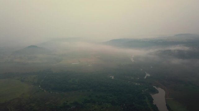 Aerial view of Gad River meandering through fog-covered agricultural valleys and hills near Malvan, Maharashtra, India during early morning. Low-lying mist blankets the rural landscape.
