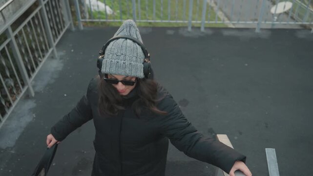Overhead shot woman descending stairs dancing in black coat and knit beanie with headphones, closeup on movement and railing grip, cinematic aerial perspective, textured concrete, deliberate rhythm,