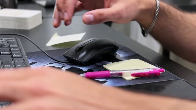 Close-up of a hand operating a computer mouse at an office desk with keyboard, sticky notes, pen, and smartphone, showing focused indoor desk work in a modern workspace.