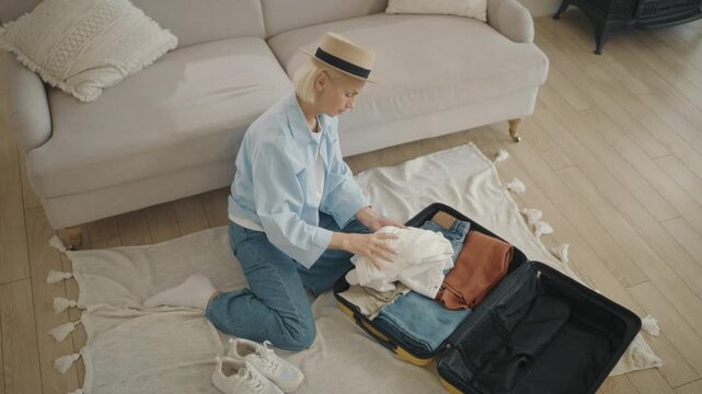 Happy elderly woman unpacking suitcase after summer trip, sitting on floor. Senior blonde lady checking clothes in travel bag, view from top, female traveler in luxury hotel room, tourism in summer
