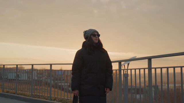 Walking woman with headphones at sunset along urban promenade, confident stride and steady rhythm, beanie and heavy coat, warm backlight and long shadows, sense of freedom and quiet empowerment,
