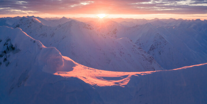 Aerial view of snow-capped mountain peaks bathed in the warm glow of the setting sun, creating a stunning contrast against the cool, blue shadows, Anaktuvuk Pass, Alaska, United States.