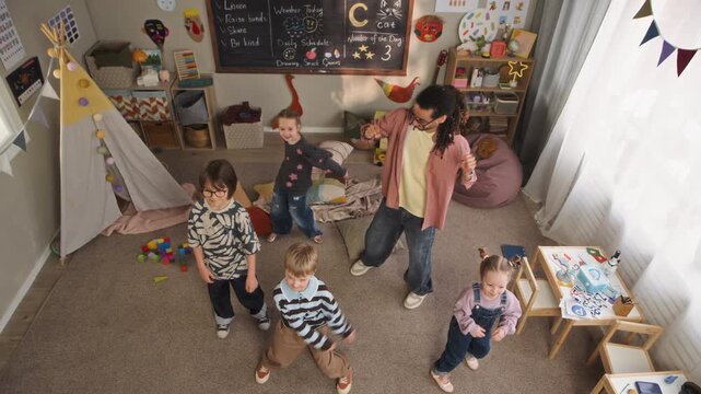 High angle full slowmo shot of cheerful young biracial male educator dancing with happy Caucasian children in cozy kindergarten classroom, having fun while sharing physical activity together