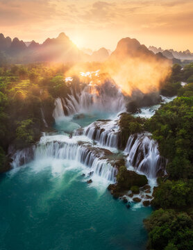 Aerial view of the majestic Ban Gioc Waterfall cascading over limestone cliffs and lush greenery under the warm glow of the setting sun, Ban Gioc Waterfall, Cao Bang, Vietnam.