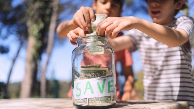 Children learning about saving money, putting dollar bills into a transparent glass jar with a save label, investing in their financial future and teaching good habits
