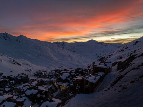 Aerial view of snow-blanketed chalets nestled in a valley, the sky ablaze with fiery oranges and soft pinks against the stark white peaks, Val Thorens, Auvergne-Rhone-Alpes, France.