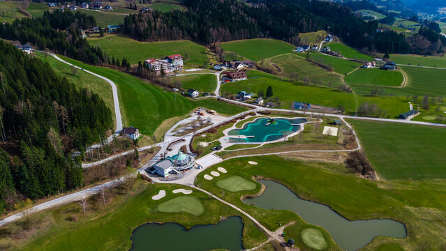 Aerial view of a vibrant green landscape dotted with buildings, a golf course, and a distinctive blue waterpark contrasting against the lush scenery, Windischgarsten, Upper Austria, Austria.