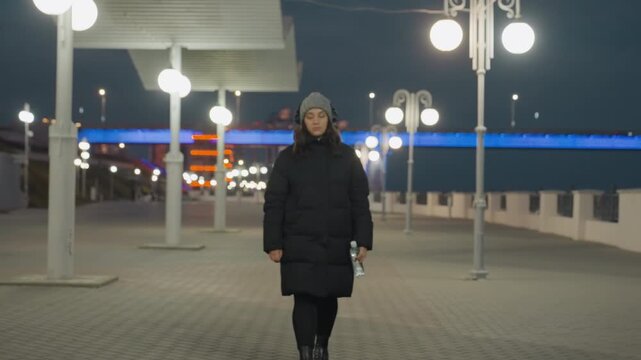 Night promenade under lamps woman dancing subtly to music, bundled in puffer coat and beanie, headphones on, empty urban walkway lined with lampposts, soft bokeh lights and cool bridge glow, slow