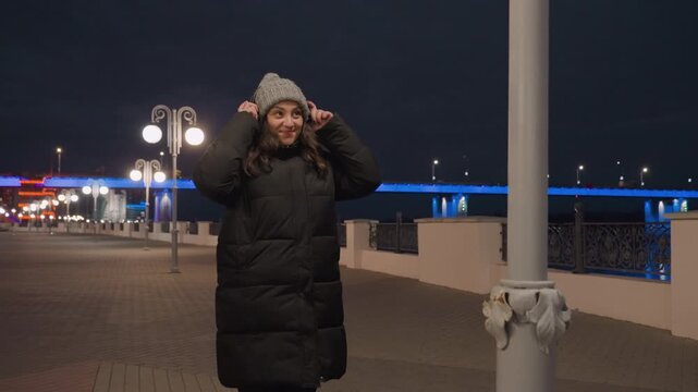 Night riverfront woman joyful dance celebration, knit hat and puffer coat, arms outstretched and laughing, spinning around lamppost, blue bridge lights and warm streetlamps, energetic urban mood