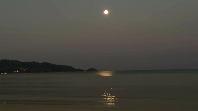 Horizontal view of a bright full moon shining high above the calm Andaman Sea water at Patong beach in Phuket, Thailand. Golden moonbeam reflection on water, scenic tropical hills dawn landscape.