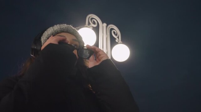 Under lamppost woman lifts sunglasses. Lowangle cinematic frames capture confident gaze and headphone beat, streetlight halo and bokeh, cool winter coat and poised model energy, slow dance gestures