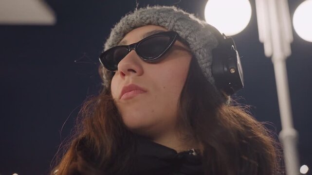 Night street headphones dance portrait showing young woman wearing sunglasses and beanie, lowangle cinematic closeup, lamplight glow, confident expression with subtle head movement, lofi city vibe