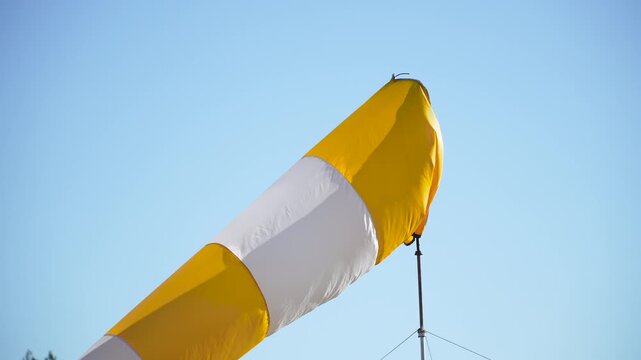 Striped windsock of white and yellow colors slowly moves against blue sky, close up. Wind sleeve shows strength and direction of wind in abstract outdoor setting