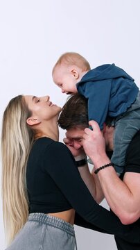 Man holding a little baby on shoulders. Woman tries to kiss her child reaching to him. White backdrop.