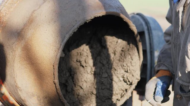 Wet Concrete Pouring From Mixer, Closeup Of Rotating Barrel Discharging Gray Slurry Into Wheelbarrow, Gloved Worker Guiding Flow With Trowel, Sunlight Casting Strong Shadows Across Textured