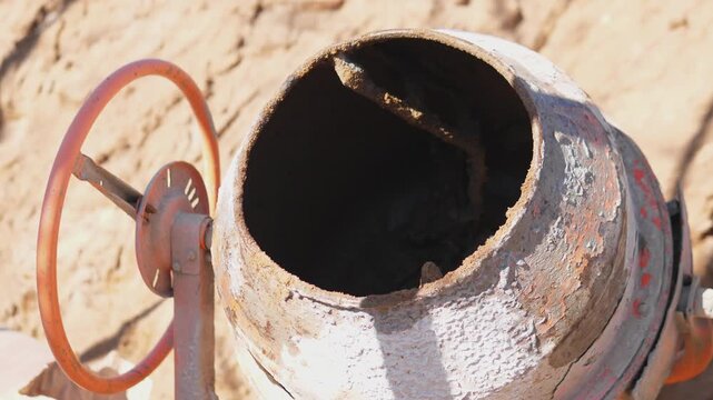 Concrete Mixer Closeup On Site, Rusted Drum And Rotating Rotor, Gloved Hand Turning Control Wheel, Sand Background, Midday Sunlight, Textured Metal And Worn Paint, Maintenance Mood,