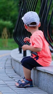 Beautiful baby boy in a cap sits on the edging near the fence. Active kid steps his feet and stands up.