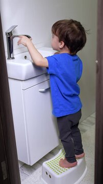 Peeping into the bathroom where little kid stands near the sink. Toddler opens and closes the tap, watching the water flow.