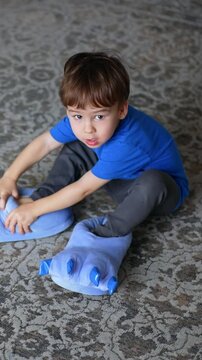 Dark-haired toddler sitting on the floor in his room. Cute child takes off his big blue soft slippers looking like claws. Top view.