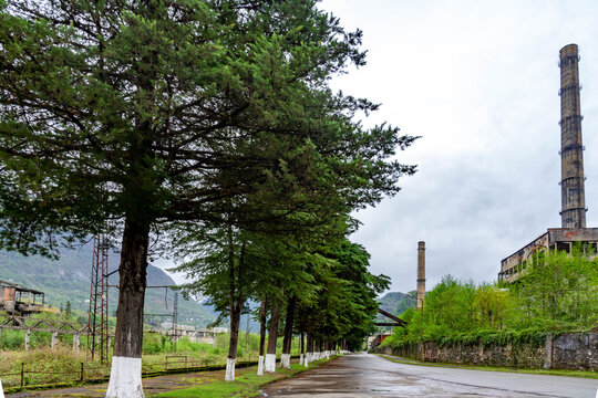 Abandoned industrial area of Tkuarchal. View of a road lined with trees leading to a ruined coal processing plant with a tall chimney under a cloudy sky in spring. Abkhazia.