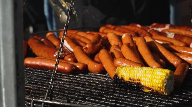 Unrecognizable person flips sausages in sunlight during outdoor grill cooking, closeup. Worker of street cafe prepares hot snack and food on grate during preparation of popular fast food.